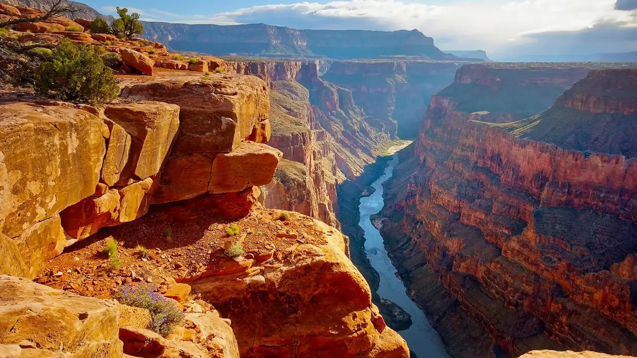 A breathtaking view of the Grand Canyon with its layered red rock cliffs and the Colorado River winding through the valley under a partly cloudy sky