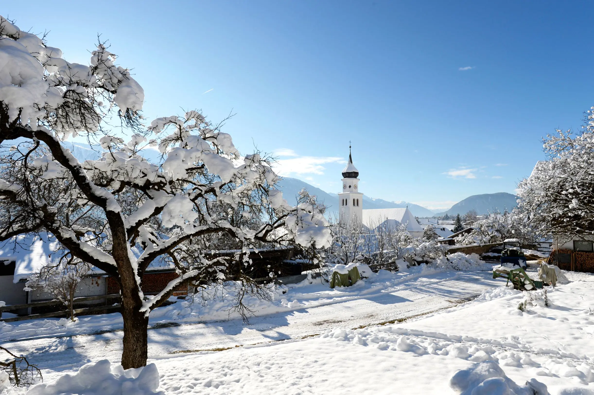 View of Seefeld village in thick snow. A leafless tree in the left forefront covered in snow, and a tall clock tower of a church slightly further away. All under a light blue sky