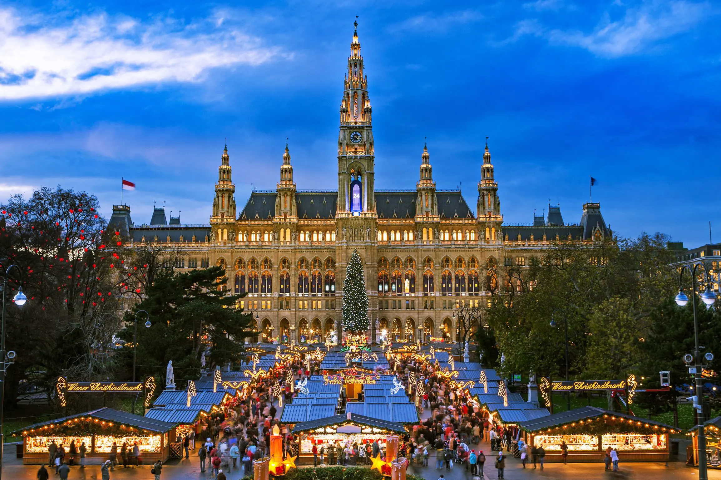 Shot of a gothic city hall which has a tall clock tower in the middle and four smaller towers, two either side, all with spiky turrets. The building has long windows all over and is a gold colour. Below are the Vienna Christmas markets, with four vertical strips of stalls, the centre having a christmas tree at the far end. There are many people shopping, with lights and lit up signs above them.