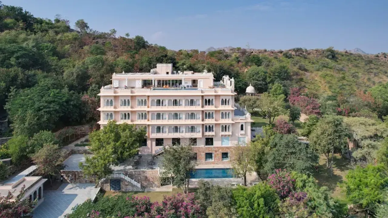 A view of Fateh Niwas Hotel in Udaipur, an elegant multi-storey heritage-style hotel surrounded by trees and hills, with balconies overlooking the landscape