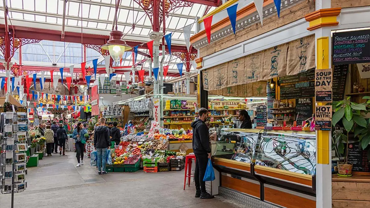 Inside view of the Jersey Market, with food stalls including one selling a  variety of vegetables in the centre, a hot food counter to the right and a rack of postcards to the left of the image. Red, white and blue banners strung across the room and a glass ceiling