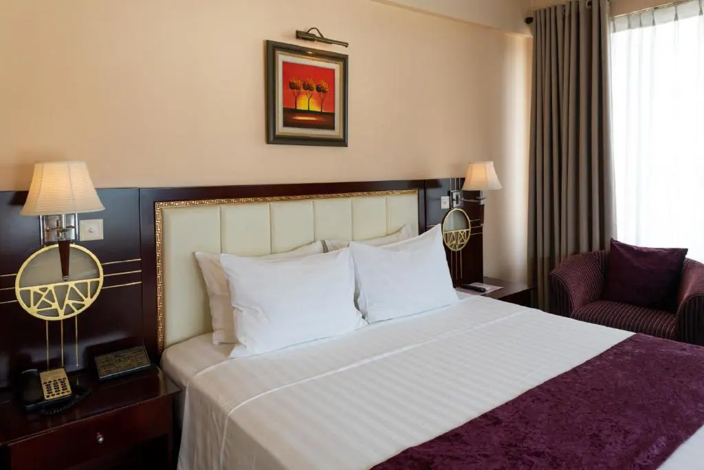 Elegant bedroom at Flower Garden Hanoi Hotel with warm wooden furniture, white linens, soft lighting, and traditional Vietnamese decor touches