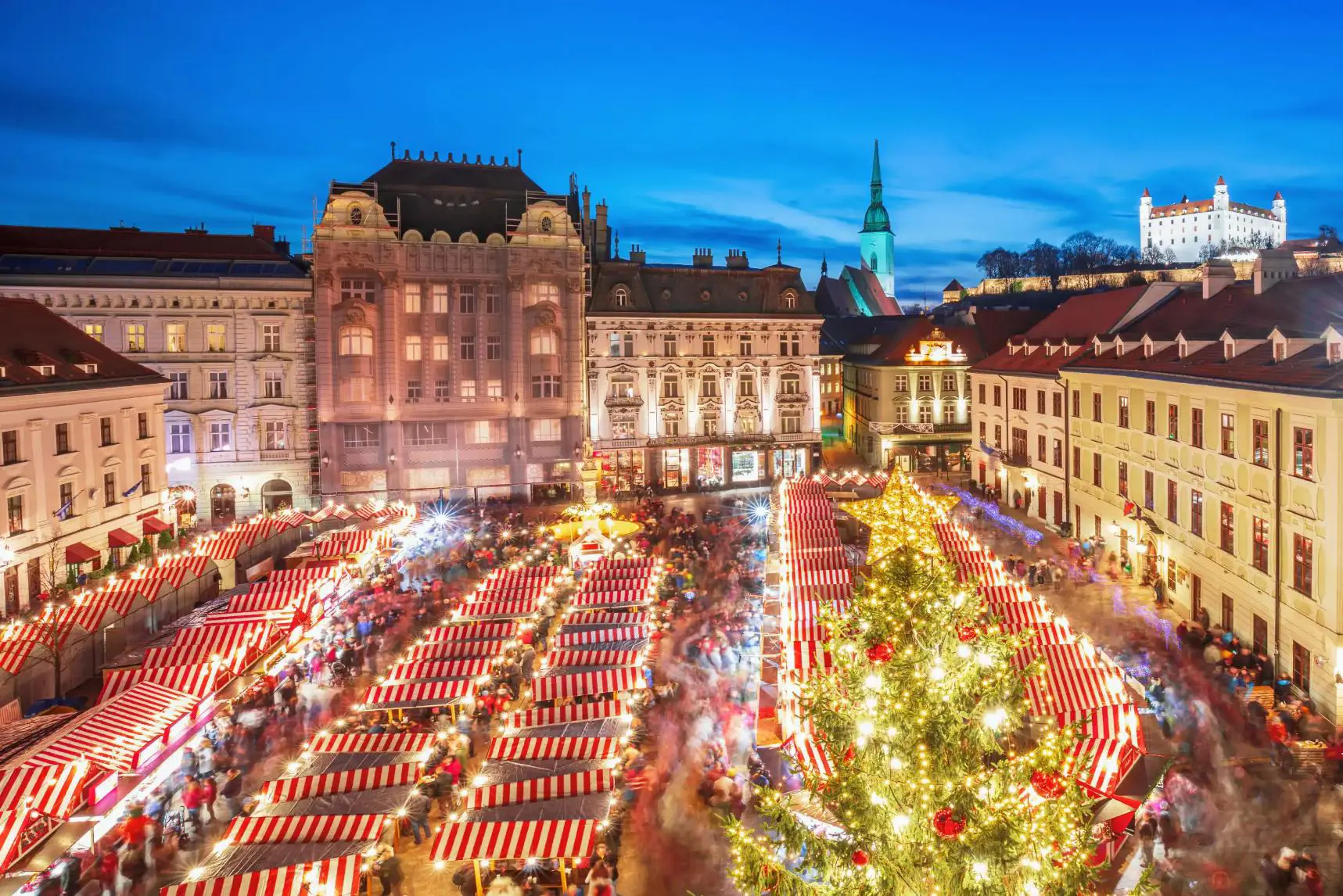 A Christmas market in Bratislava's main square, featuring festive wooden stalls decorated with lights, a large Christmas tree, and crowds of people enjoying the festive atmosphere