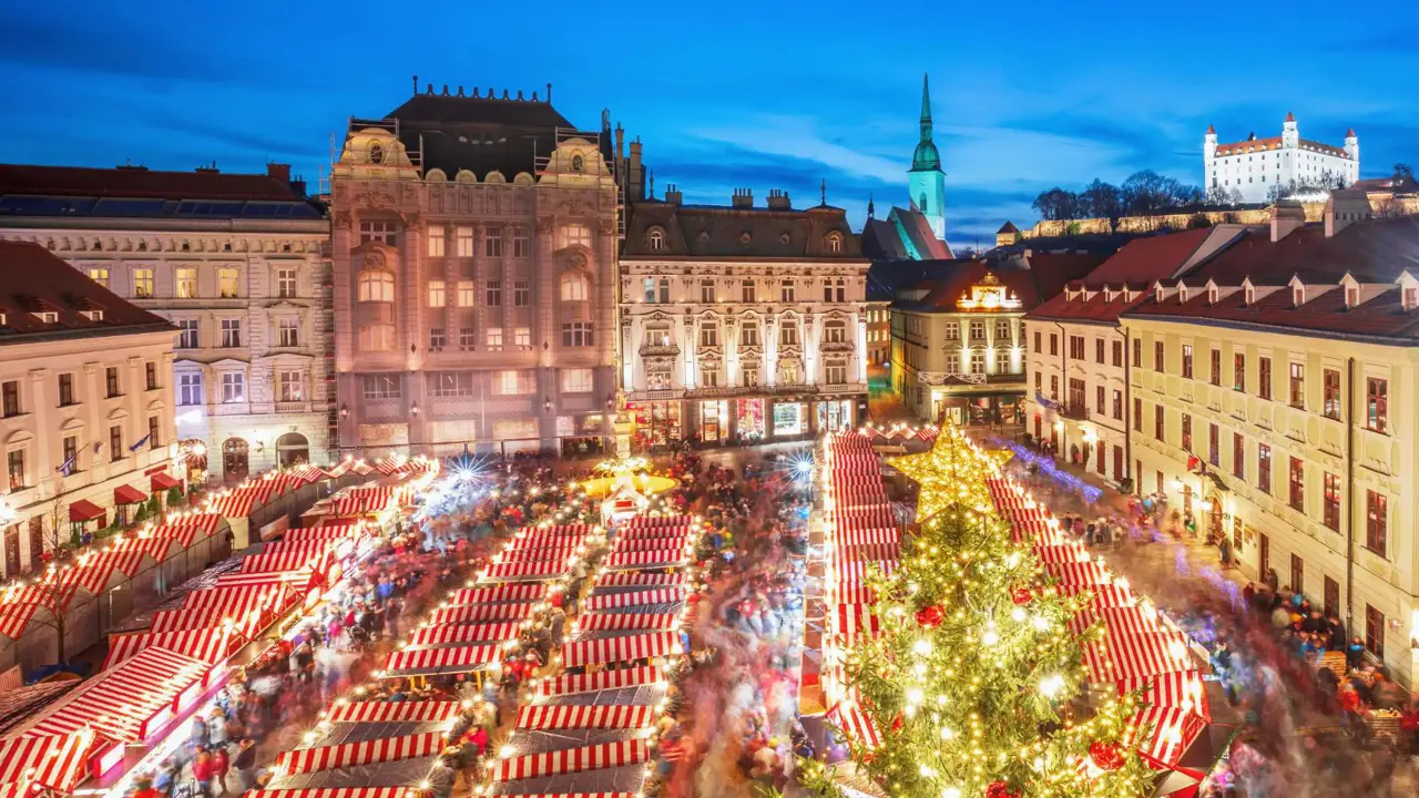 A Christmas market in Bratislava's main square, featuring festive wooden stalls decorated with lights, a large Christmas tree, and crowds of people enjoying the festive atmosphere