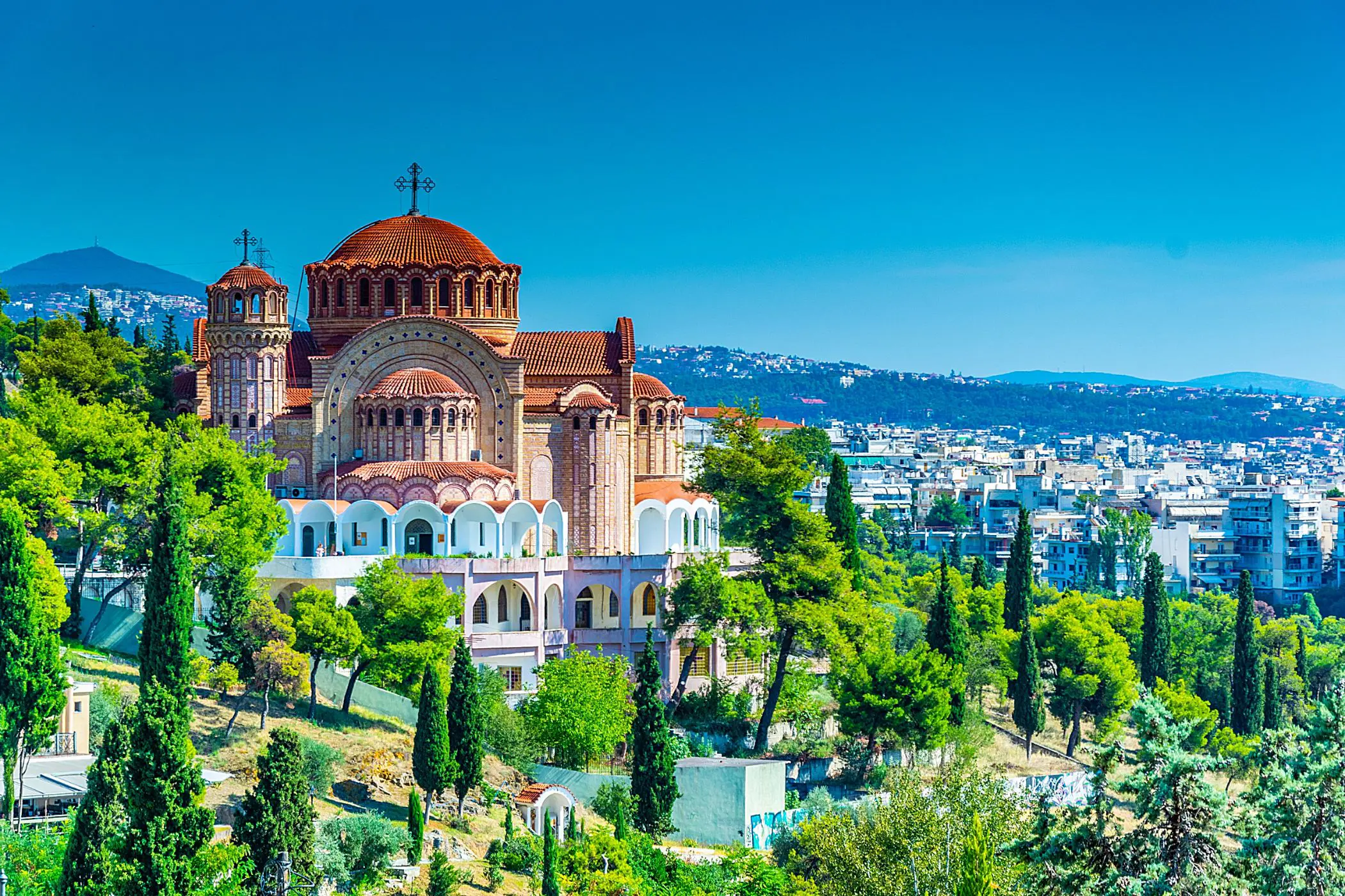 Brown and beige cathedral with a domed top with a cross on, trees in the forefront and a view of a town in the distance