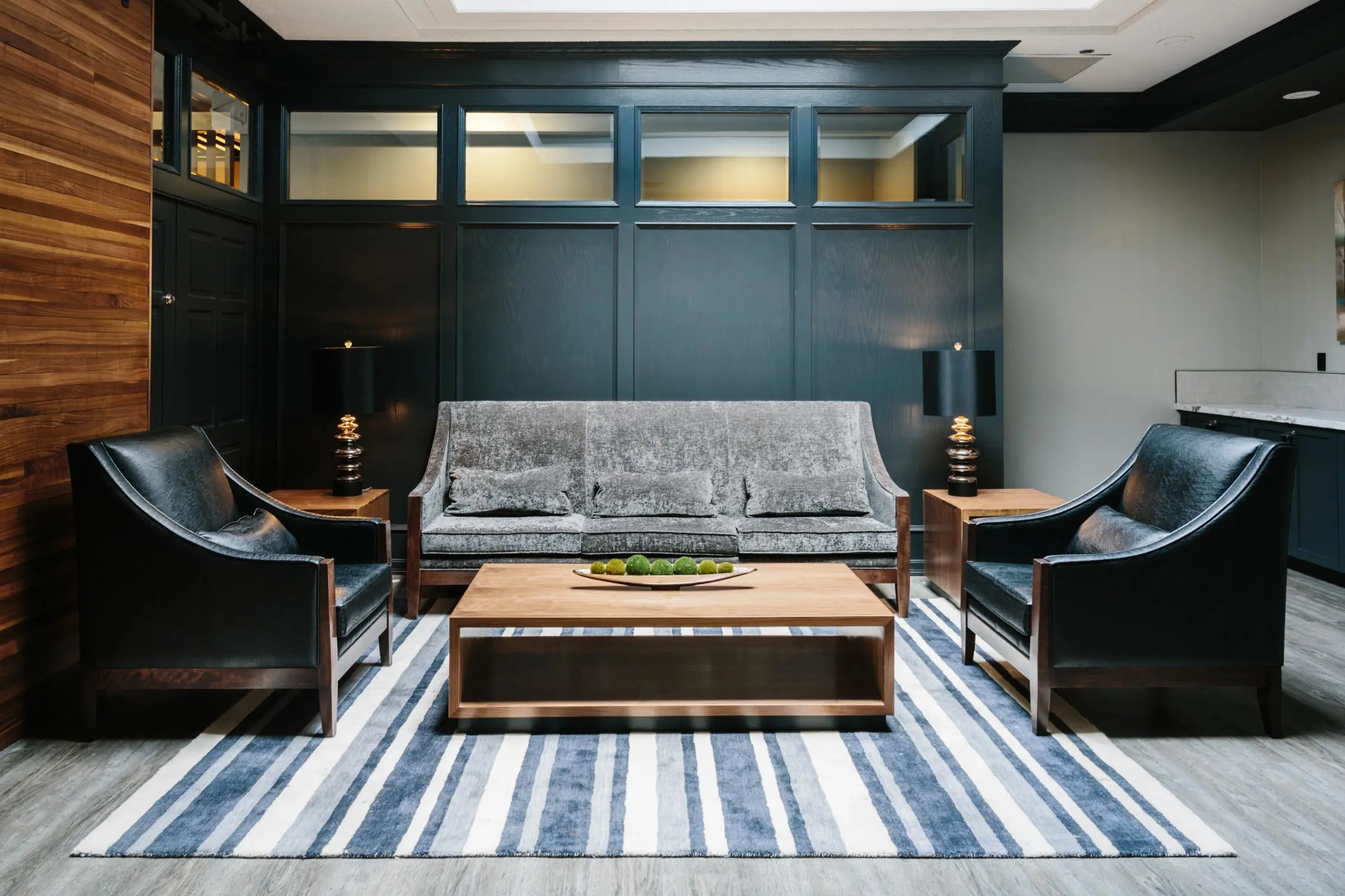Cosy lounge at Elk + Avenue hotel with a velvet grey sofa, black leather armchairs, a wooden coffee table on a striped rug, and soft lighting set against dark wood paneling