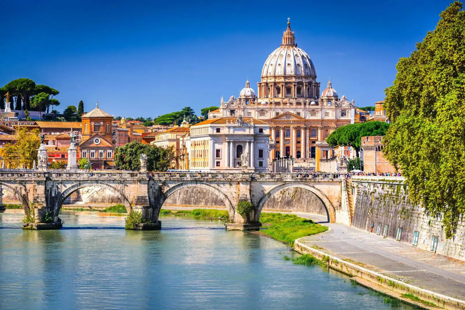 View of St Peter's Basilica in Vatican City, with the River Tiber and the historic Ponte Sant'Angelo bridge in the foreground, on a bright sunny day in Rome, Italy