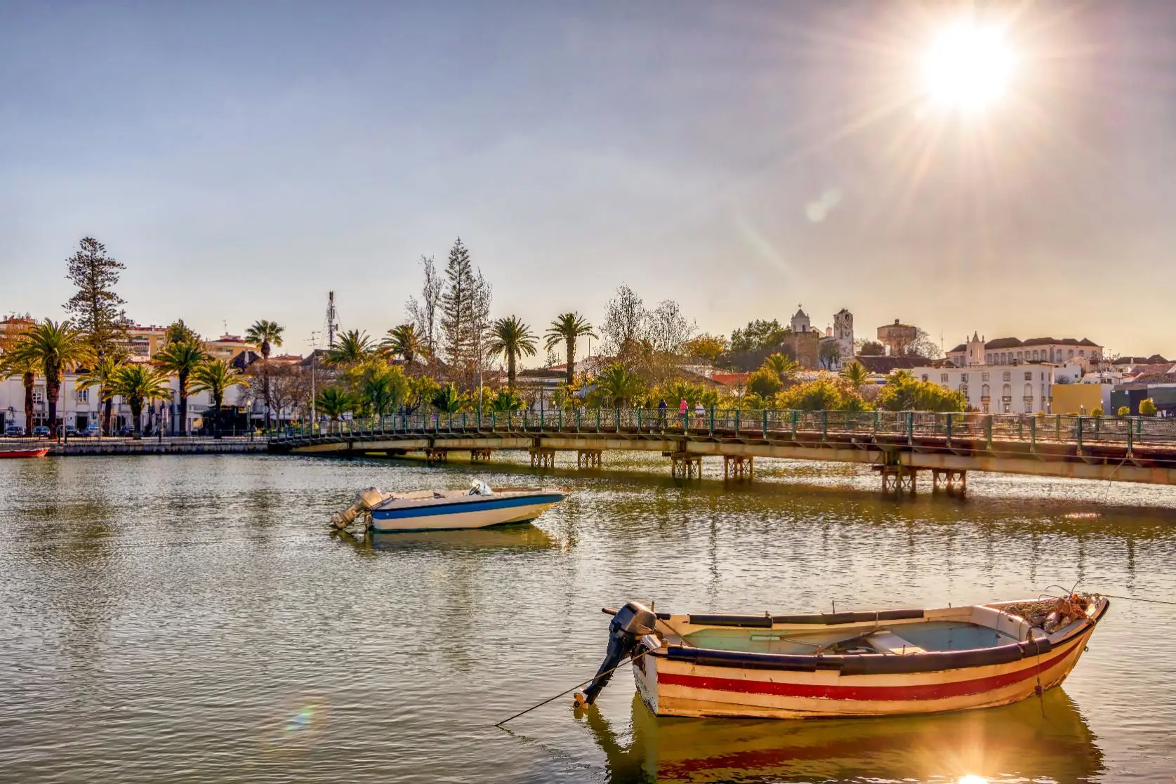 Two small boats moored in front of a bridge in Tavira on the Algarve, with trees and buildings lining the water’s edge in the background