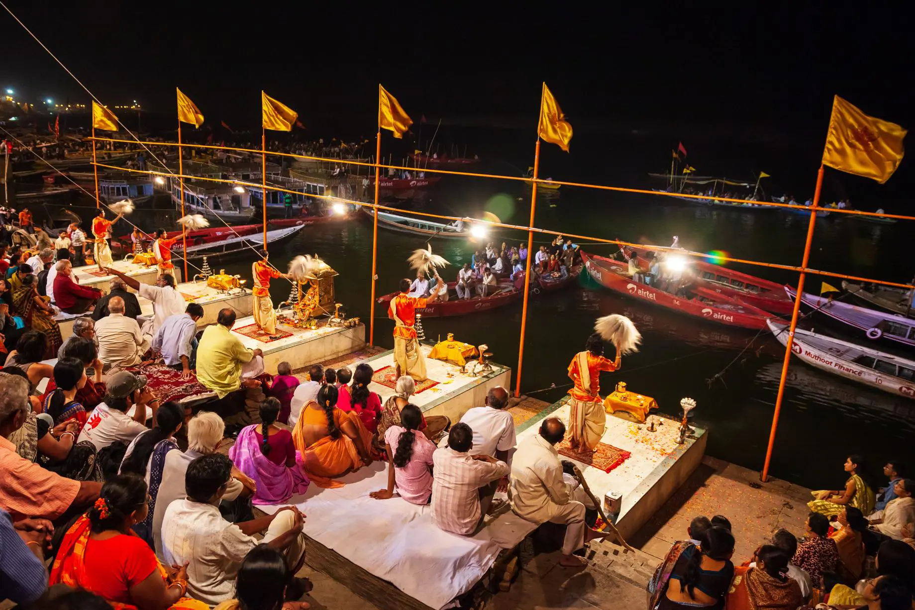 Priests dressed in traditional saffron robes performing the Ganga Aarti ceremony on the ghats of Varanasi at night, surrounded by devotees and illuminated by rows of lights along the riverbank