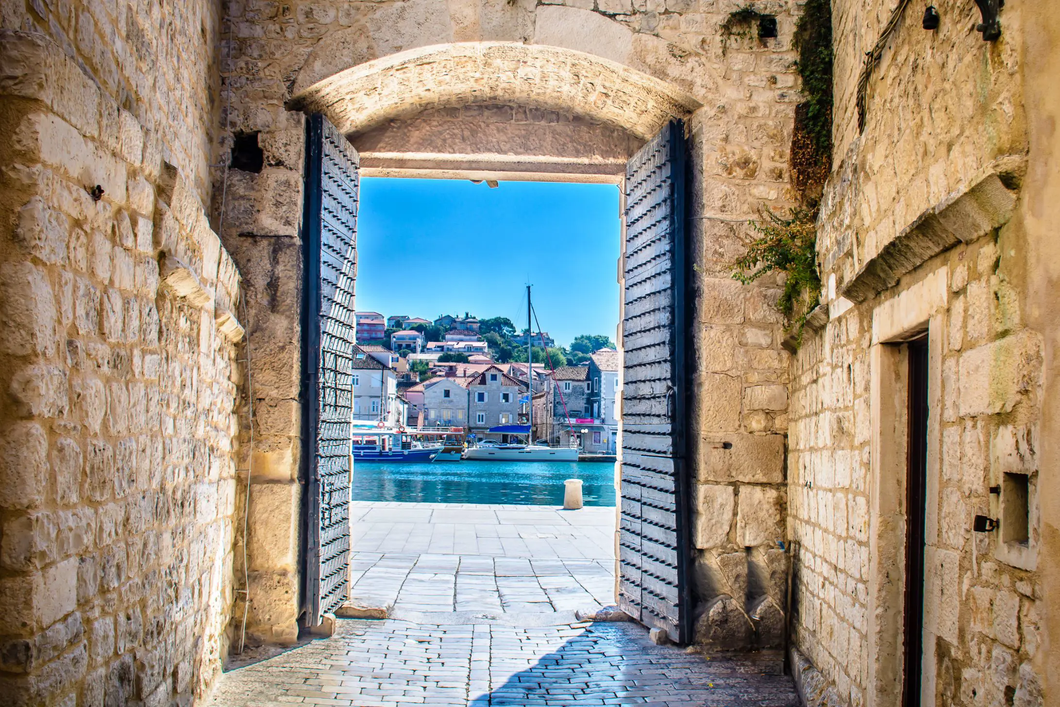 View of a boat on some water, with buildings behind it, through open doors in a stone walkway. A bright blue sky.