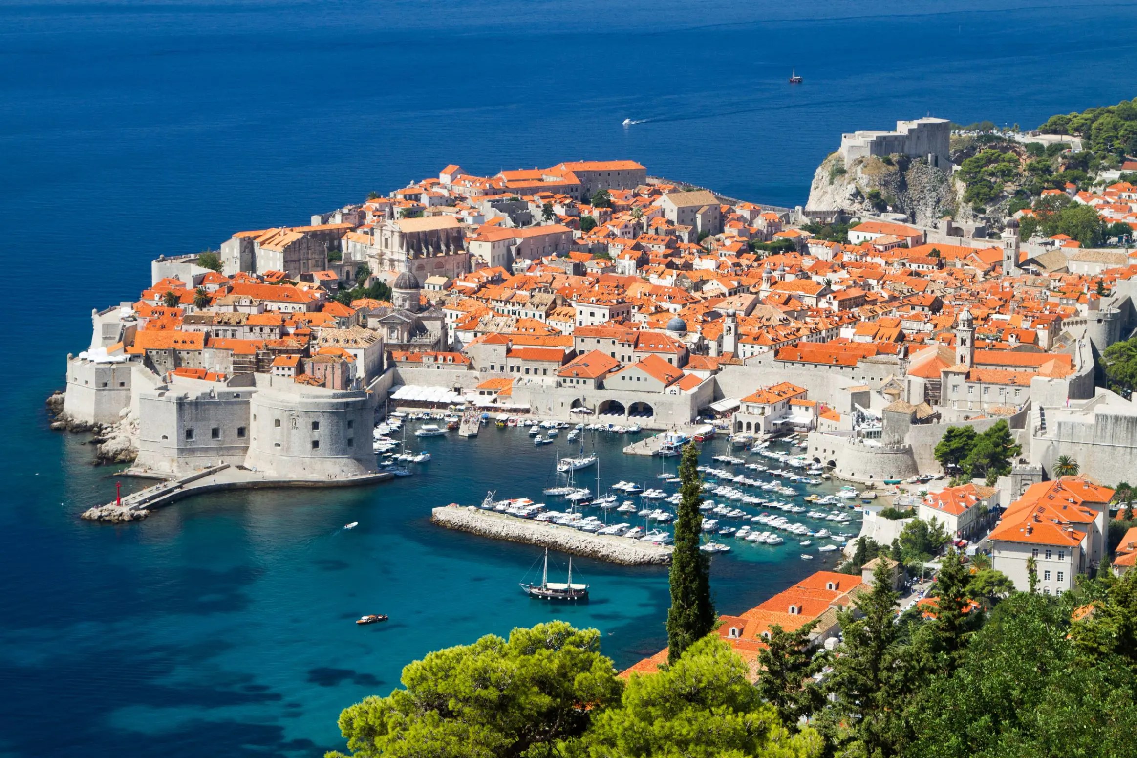 High angle view of Dubrovnik's coast and its harbour, with boats docked and lots of buildings with orange roofs behind. The navy blue sea to the left.