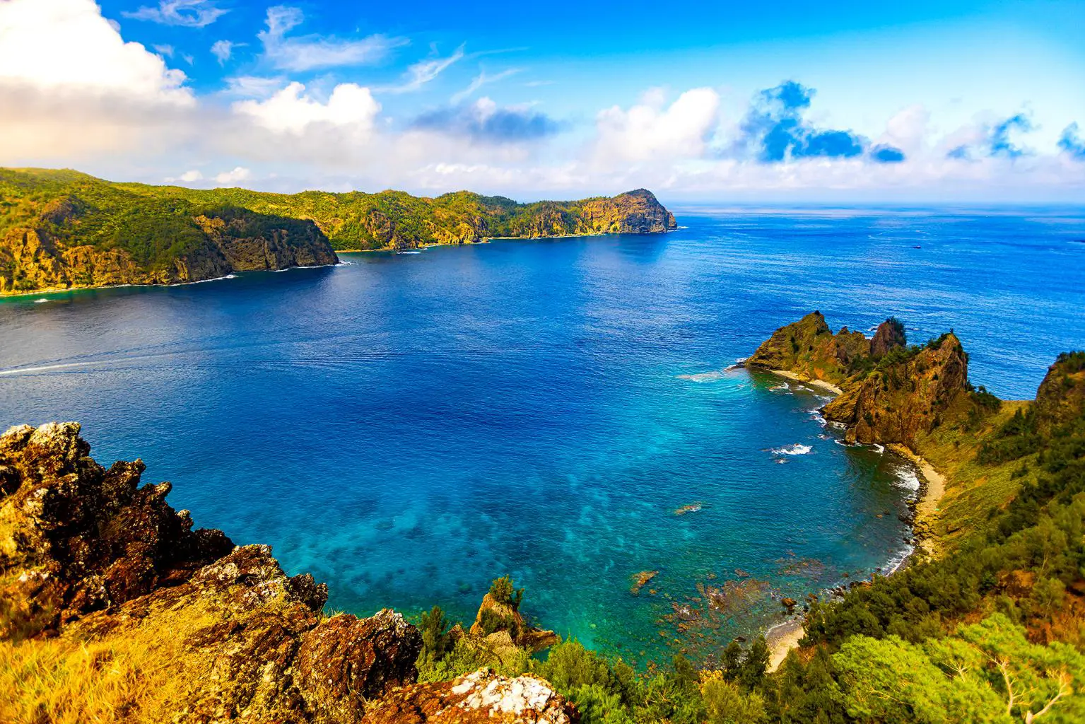 A stunning coastal view from Nagasaki Observatory on Chichijima Island, with clear blue waters, rugged cliffs, and lush green hills under a vibrant sky