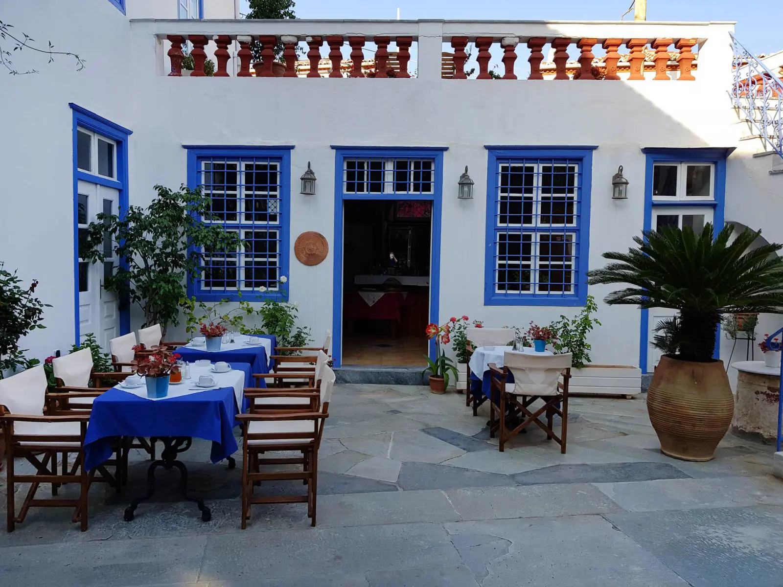 Outdoor dining area on a patio at Hotel Ippokampos in Hydra, Greece, featuring tables with blue tablecloths, surrounded by potted plants, with doors and windows framed in traditional blue paint