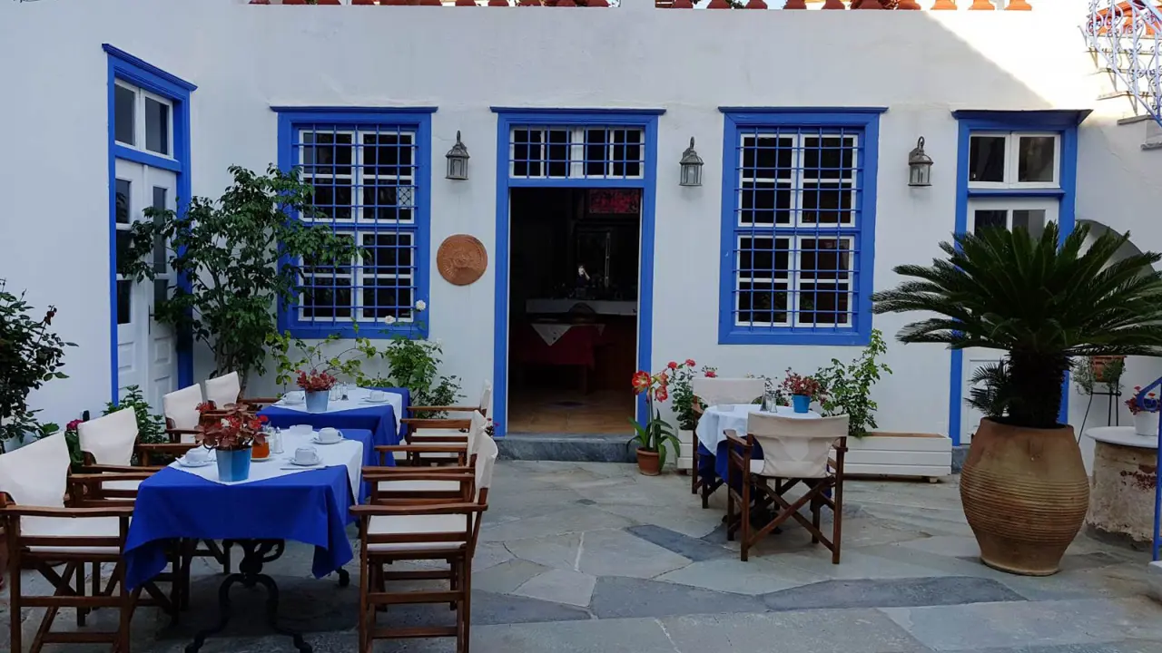 Outdoor dining area on a patio at Hotel Ippokampos in Hydra, Greece, featuring tables with blue tablecloths, surrounded by potted plants, with doors and windows framed in traditional blue paint