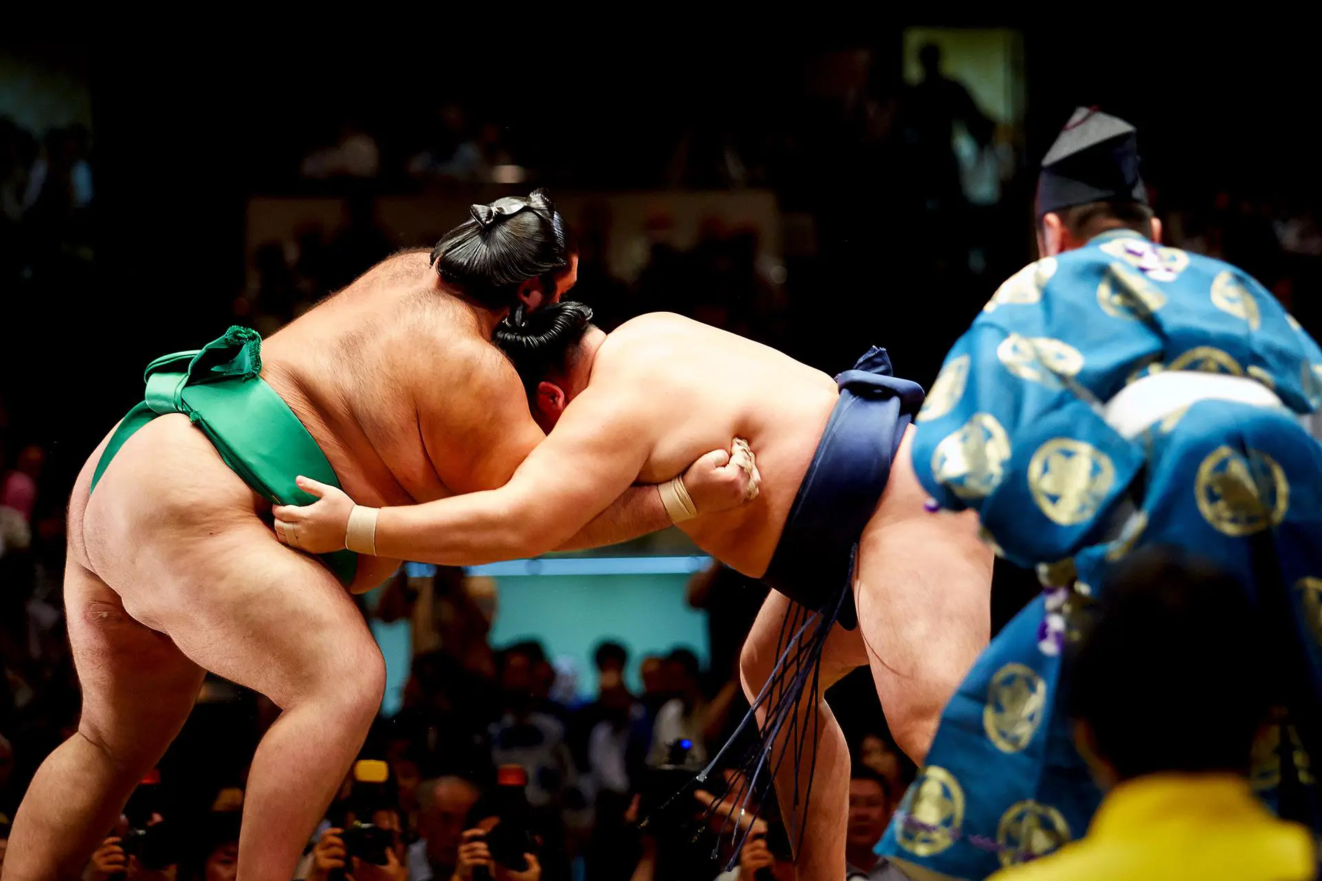 Close-up of sumo wrestlers locked in action during a bout, surrounded by a referee (gyōji) and onlookers in a sumo arena in Fukuoka