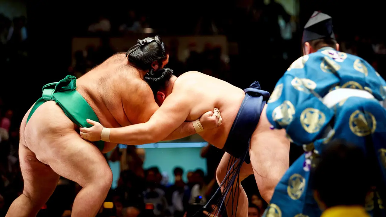 Close-up of sumo wrestlers locked in action during a bout, surrounded by a referee (gyōji) and onlookers in a sumo arena in Fukuoka