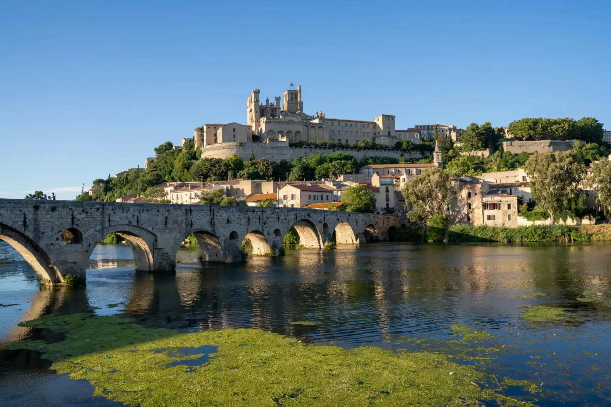 A historic stone bridge crossing a river, leading to the ancient town of Narbonne in southern France, with old buildings and a large cathedral on a hill under a clear blue sky