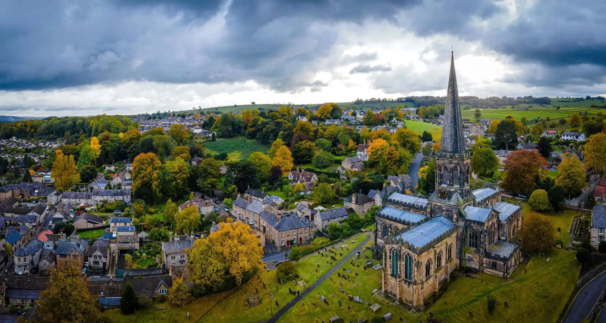 An aerial view of Bakewell in the Peak District, England, with a tall church spire, stone cottages, colourful autumn trees, and rolling green hills under a dramatic cloudy sky