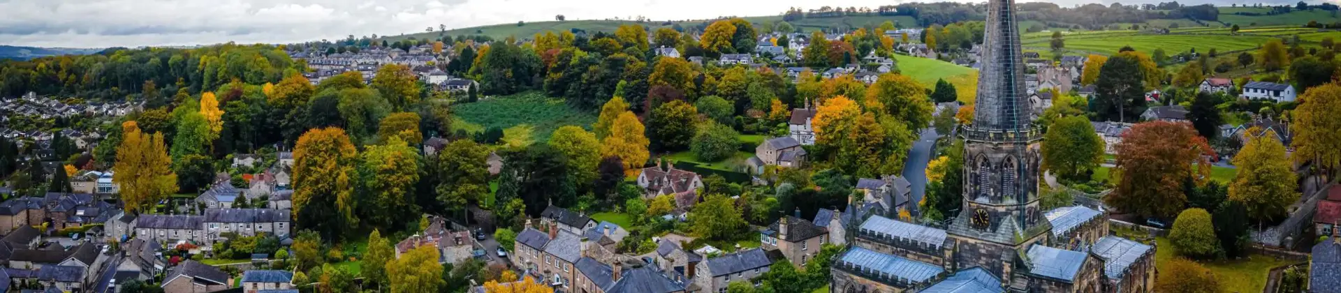 An aerial view of Bakewell in the Peak District, England, with a tall church spire, stone cottages, colourful autumn trees, and rolling green hills under a dramatic cloudy sky