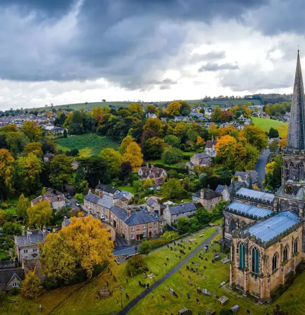 An aerial view of Bakewell in the Peak District, England, with a tall church spire, stone cottages, colourful autumn trees, and rolling green hills under a dramatic cloudy sky