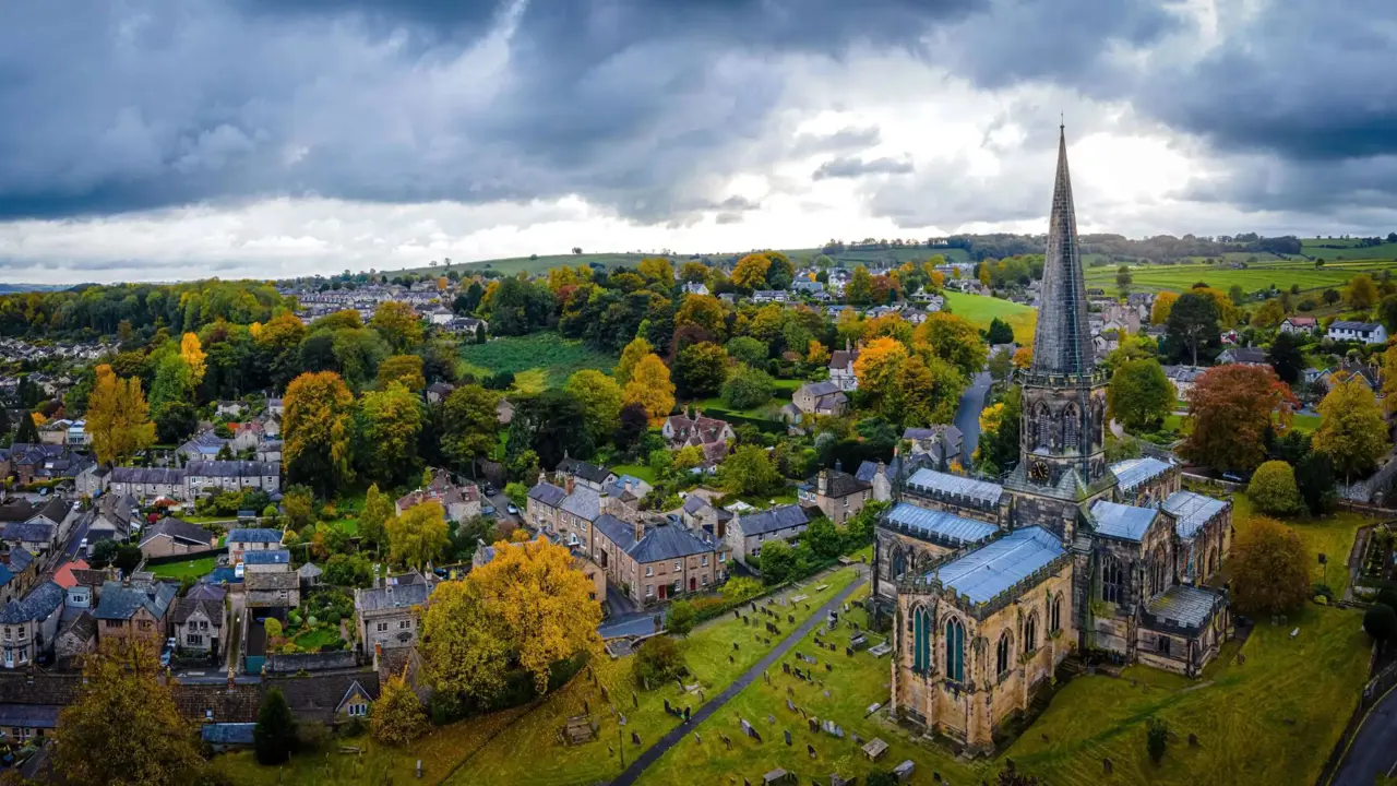 An aerial view of Bakewell in the Peak District, England, with a tall church spire, stone cottages, colourful autumn trees, and rolling green hills under a dramatic cloudy sky