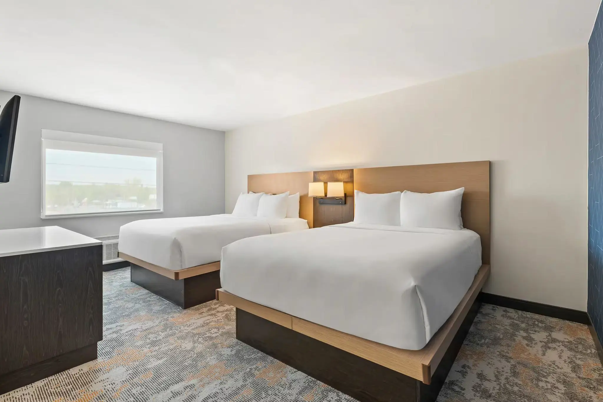 Hotel suite featuring two queen-sized beds with white bedding, a small sitting area, modern furnishings, and a large window letting in daylight