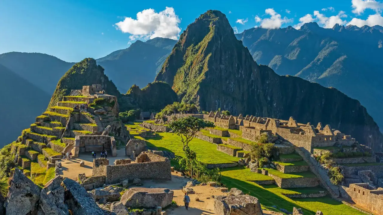 A panoramic view of Machu Picchu in Peru, showing ancient Incan ruins built into a green mountain ridge under a clear blue sky, with Huayna Picchu towering in the background