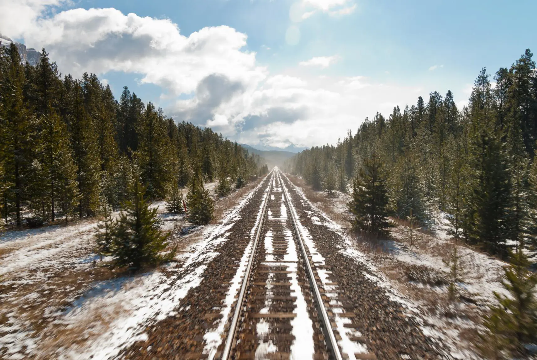 Snow-covered railway tracks extending through a wintry Canadian landscape, bordered by tall green trees on both sides