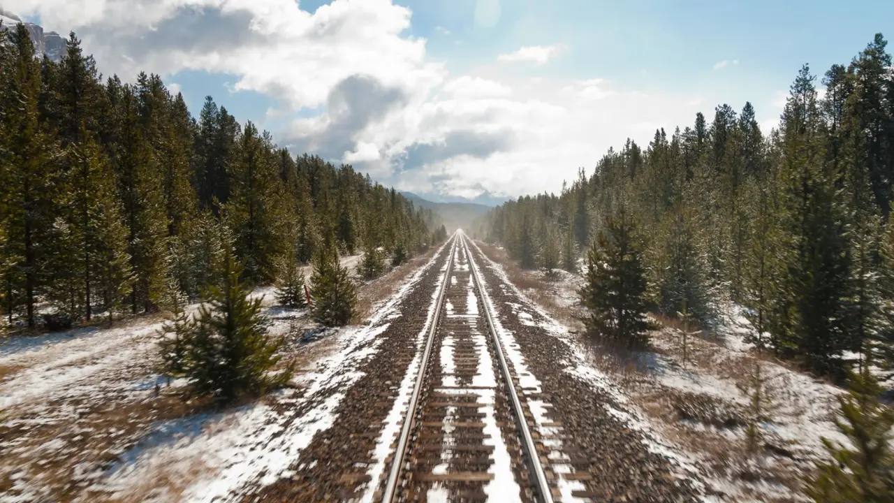 Snow-covered railway tracks extending through a wintry Canadian landscape, bordered by tall green trees on both sides