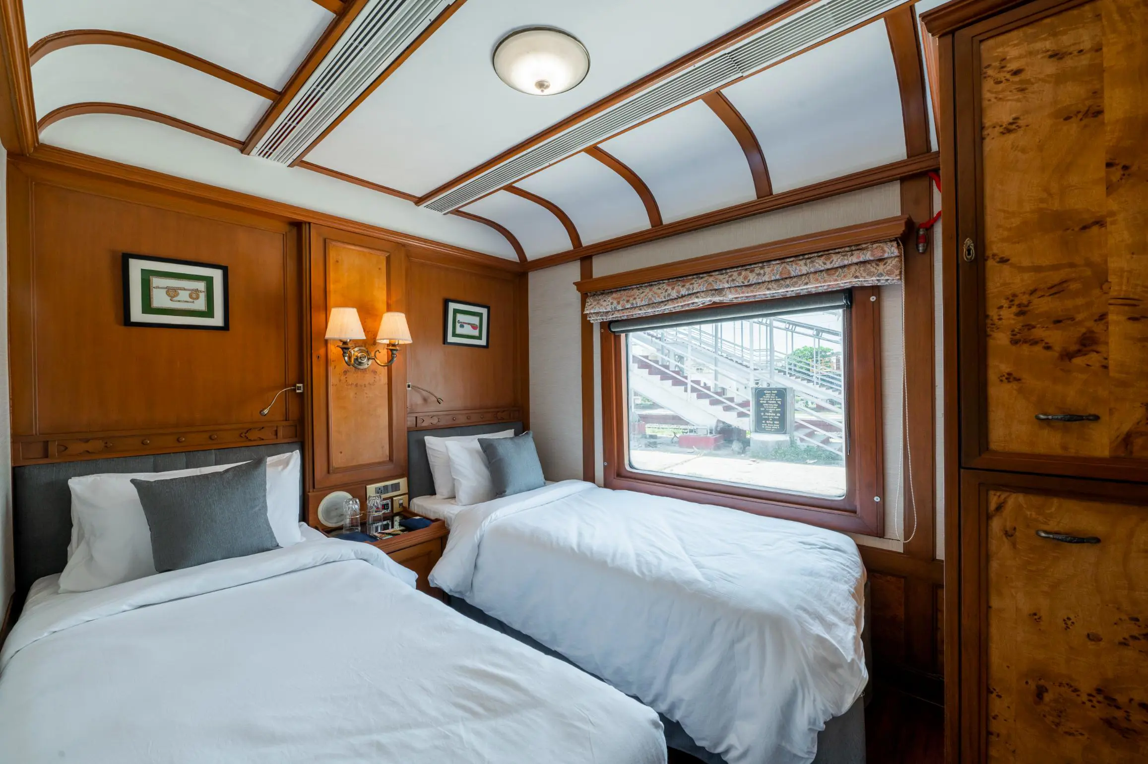 Interior of a Deccan Odyssey twin room, showing two neatly made beds, elegant wooden furnishings, and a large window with a patterned blind
