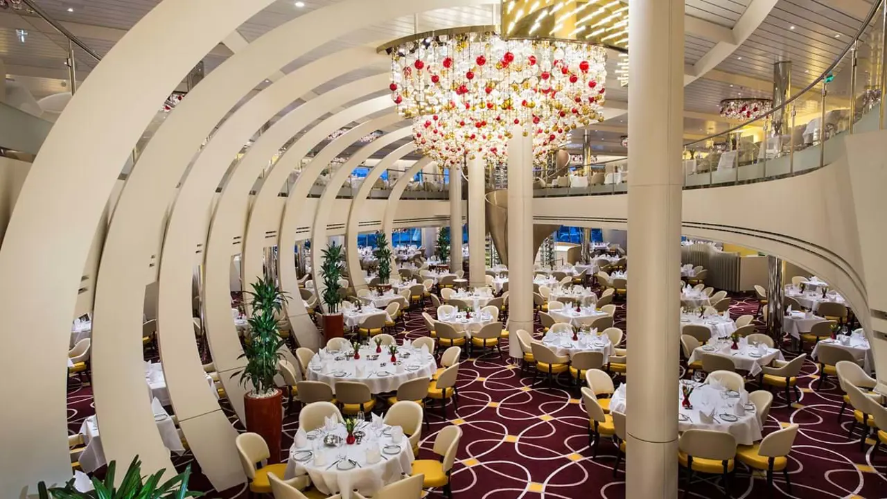 The elegant dining room on board MS Koningsdam, featuring a large chandelier, gold decor, and neatly set tables with comfortable seating