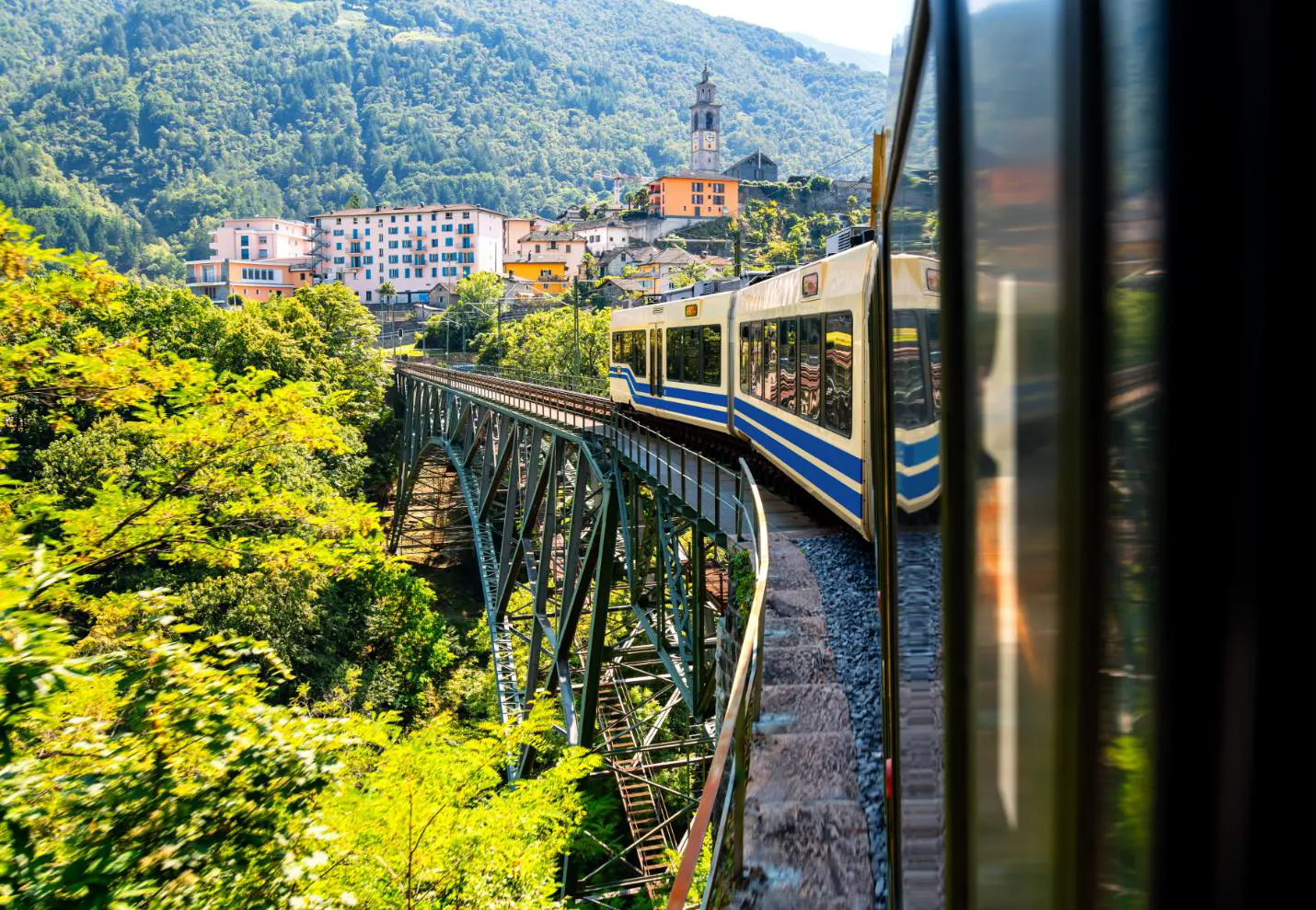 Centovalli Railway bridge in Switzerland with a train crossing, surrounded by mountainous countryside, and a town visible in the distance