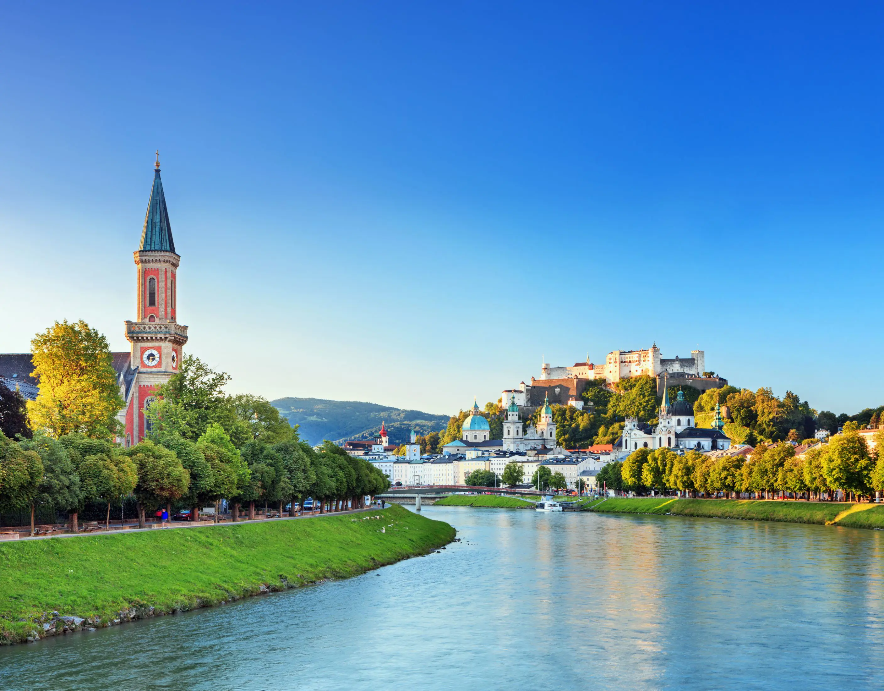 View of a river winding to the left, with grass and trees on either side, and a red church with a tall, slim tower with a blue turret. On the left of the river multiple large buildings can be seen, with numerous turquoise turrets pointing out, and the fortress on the top of the hill, all under a blue sky