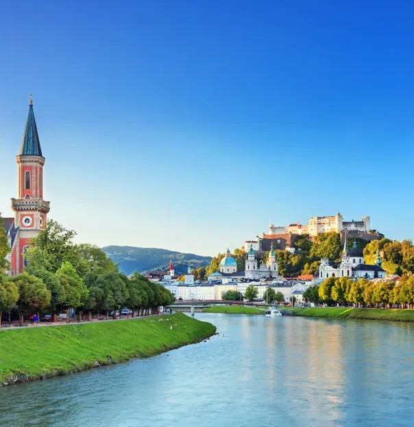 View of a river winding to the left, with grass and trees on either side, and a red church with a tall, slim tower with a blue turret. On the left of the river multiple large buildings can be seen, with numerous turquoise turrets pointing out, and the fortress on the top of the hill, all under a blue sky