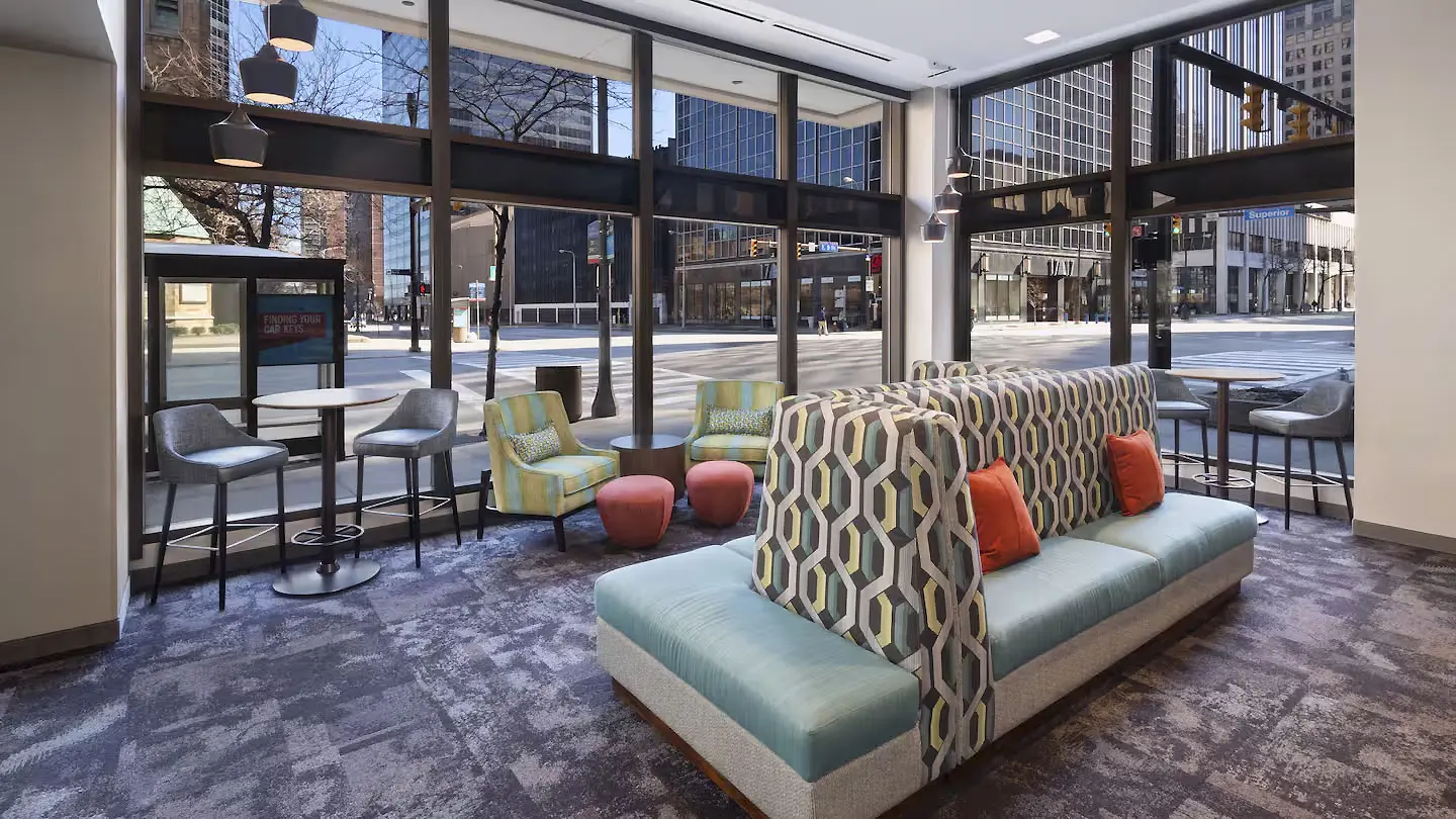 The lounge at Hampton Inn Downtown, Cleveland, featuring contemporary seating in the centre with green upholstery and deep orange cushions. There are large street-facing windows that fill the space with natural light