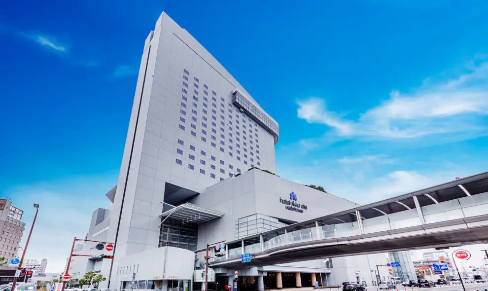 Exterior of Hotel Nikko Ōita Oasis Tower, a tall, modern building with a skybridge connecting to the main entrance and a bright blue sky overhead