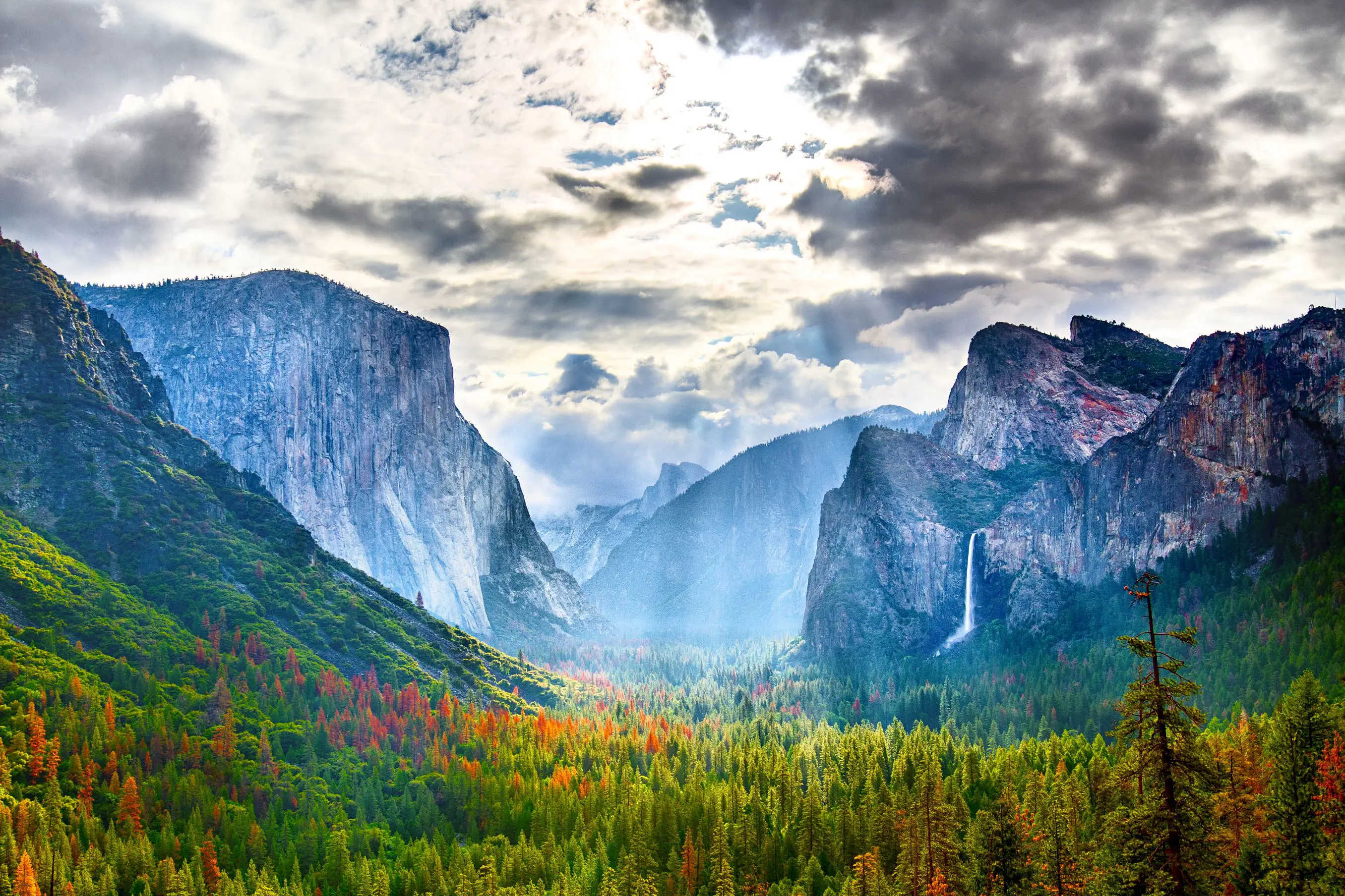 Yosemite National Park in California with towering granite cliffs, green forested areas, and cloudy skies with sun rays breaking through