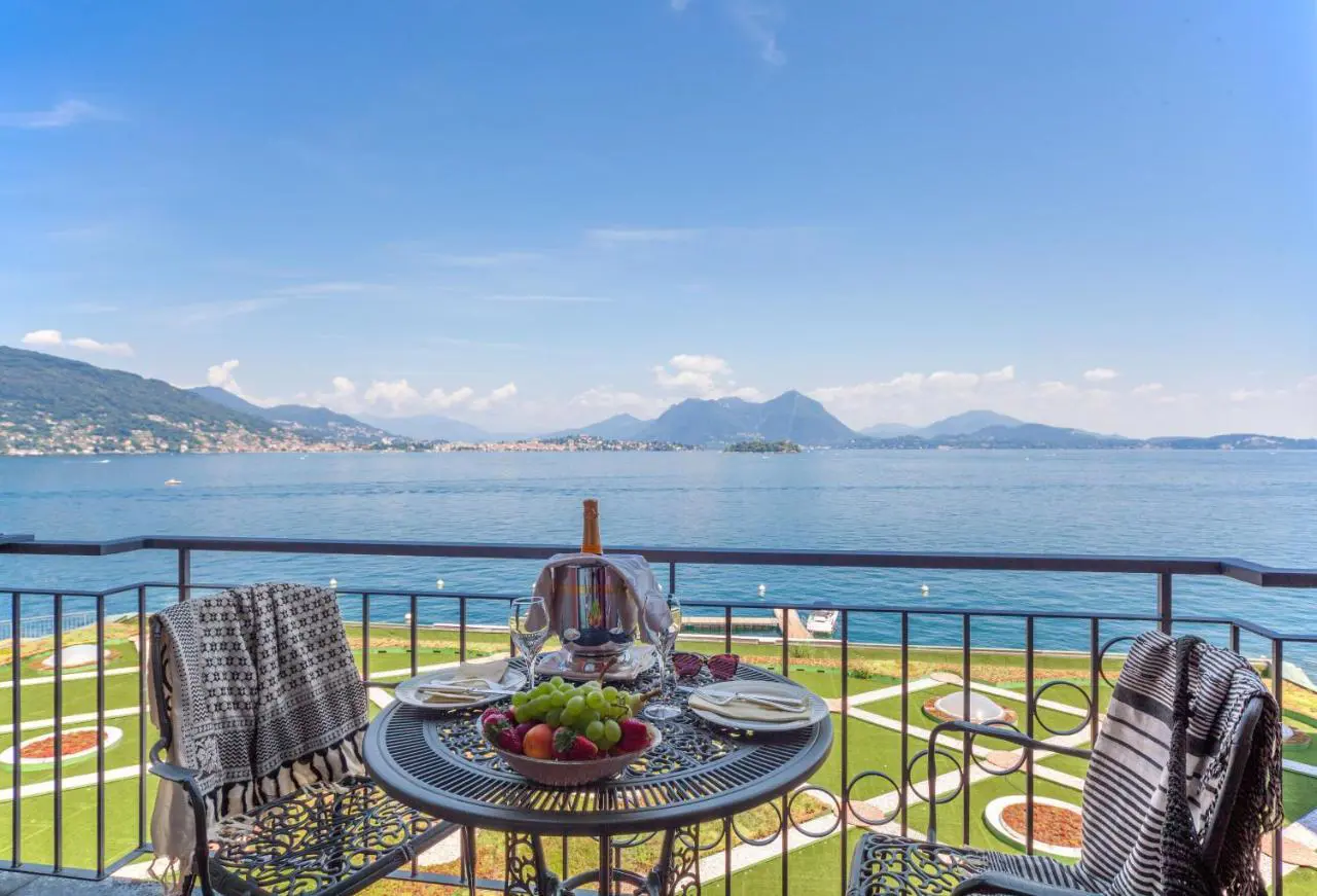 A table with two chairs on a balcony at the Grand Hotel Dino in Baveno, set with fruit and two plates, overlooking the lake, mountains, and landscaped gardens below