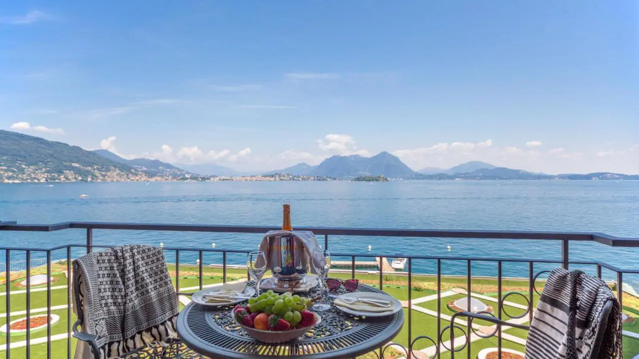 A table with two chairs on a balcony at the Grand Hotel Dino in Baveno, set with fruit and two plates, overlooking the lake, mountains, and landscaped gardens below