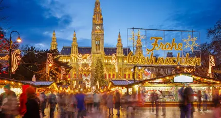 Shot of Vienna's gothic city hall which has one tall clock tower and four smaller towers, two on each side, all with pointy turrets. The building is a gold colour and is in front of a bright blue night sky. In front, is a Christmas Market, showing strips of cabin-like stalls and a variety of lit up Christmas decorations, one in the forefront reading 'Merry Christmas' in Austrian. People can be seen, although blurry as they are moving around. 