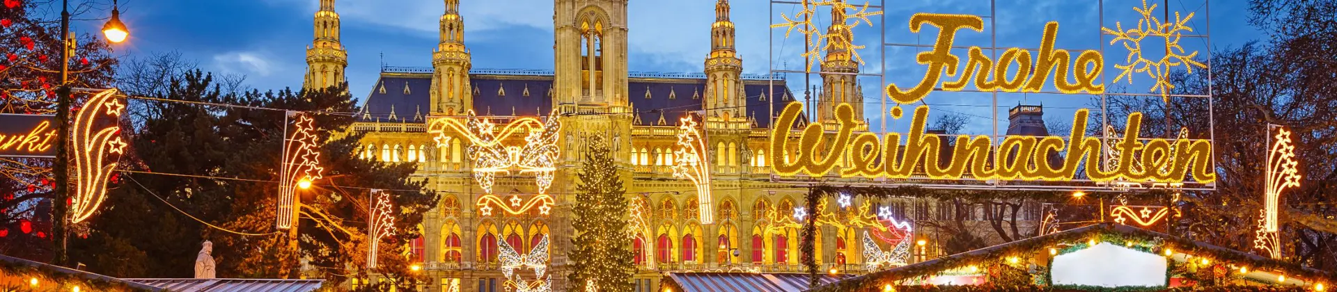Shot of Vienna's gothic city hall which has one tall clock tower and four smaller towers, two on each side, all with pointy turrets. The building is a gold colour and is in front of a bright blue night sky. In front, is a Christmas Market, showing strips of cabin-like stalls and a variety of lit up Christmas decorations, one in the forefront reading 'Merry Christmas' in Austrian. People can be seen, although blurry as they are moving around. 
