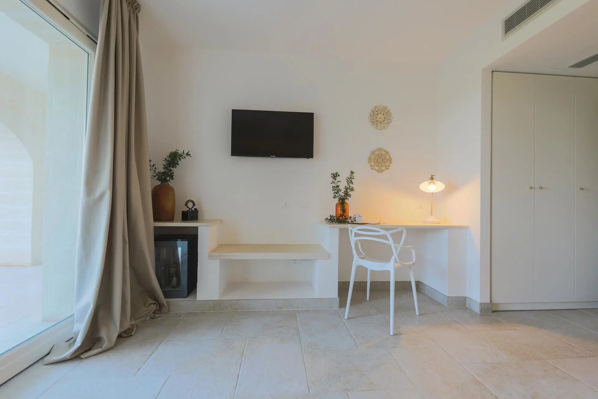 Bright minimalist room at Masseria Casselli Hotel featuring a wall-mounted TV, white desk and chair, a table lamp, decorative plants, and beige curtains covering a large glass door
