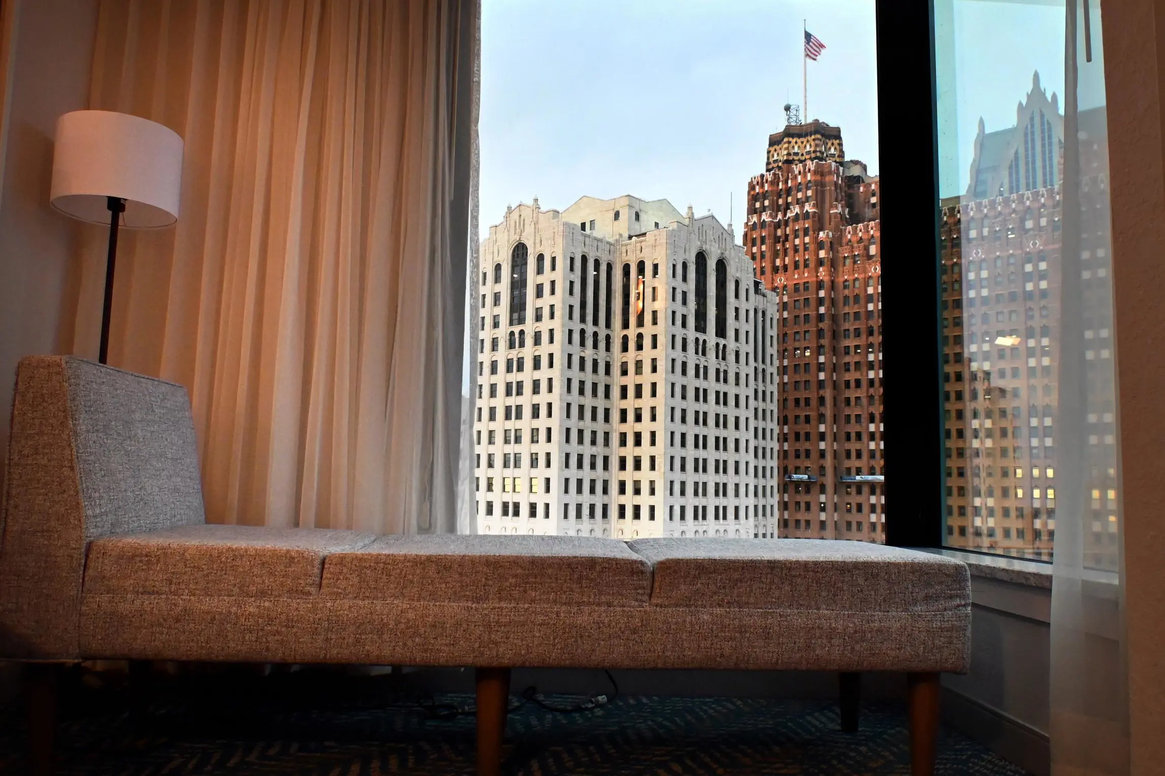 City view from a standard room at Fort Pontchartrain hotel, featuring a modern grey chaise lounge, floor lamp, and large windows overlooking historic high-rise buildings in downtown Detroit