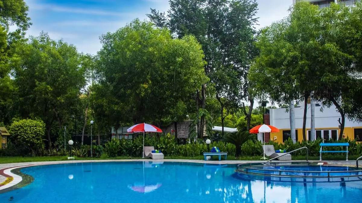 Exterior view of the pool at the Fortune Pandiyan Hotel in Madurai, framed by tall trees and greenery, with sun loungers and red-and-white striped parasols around it