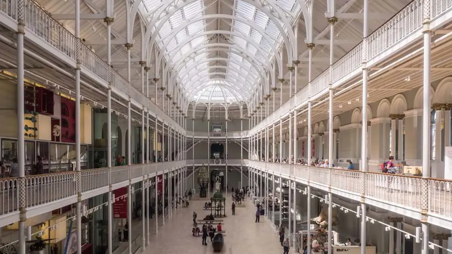 Inside the National Museum of Scotland, showing the glass ceiling and white railings of the different floor levels on either side, and people stood on the ground floor