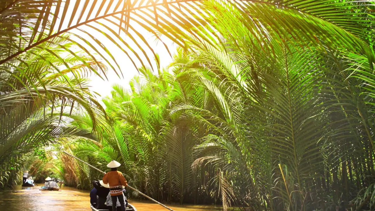 Small wooden boats glide through a palm-lined canal off the Mekong River in Thailand, with a person in a conical hat steering under filtered sunlight