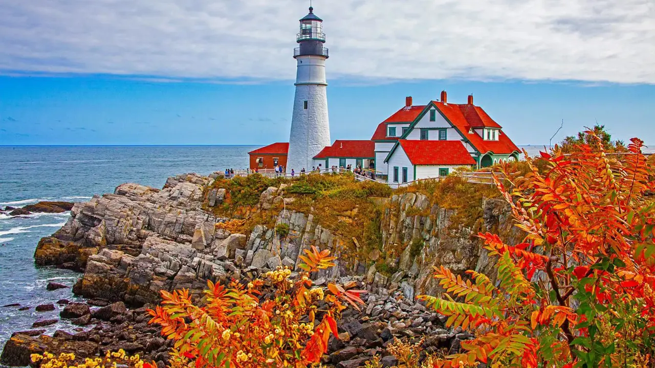 White lighthouse and red-roofed buildings on a rocky coast in Portland, with autumn foliage in the foreground