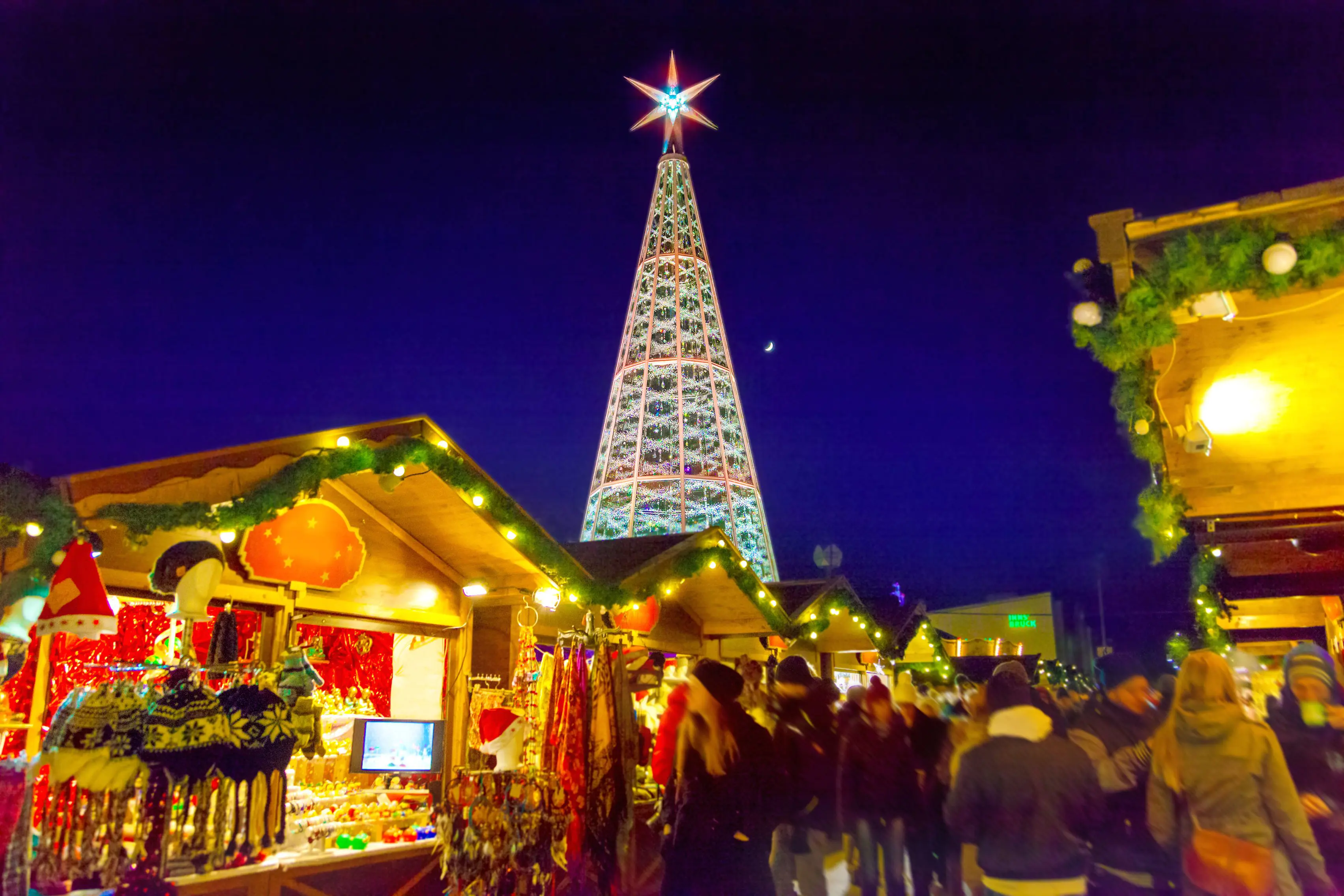 View of a Christmas market, with wooden, cabin-like stalls in the forefront, selling winter hats, dream catchers and scarves. The roofs are lined with green leaf-like garlands and fairy lights. Behind these is a Christmas tree metal structure, with a large, lit up star on top, in front of a navy blue night sky.