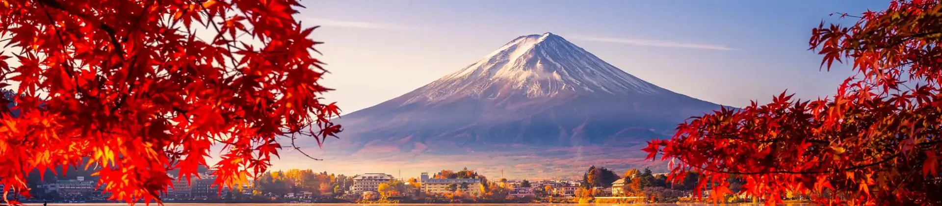 Mount Fuji reflected in Lake Kawaguchi, framed by vivid red autumn leaves, with a sunlit lakeside town between the water and the mountain