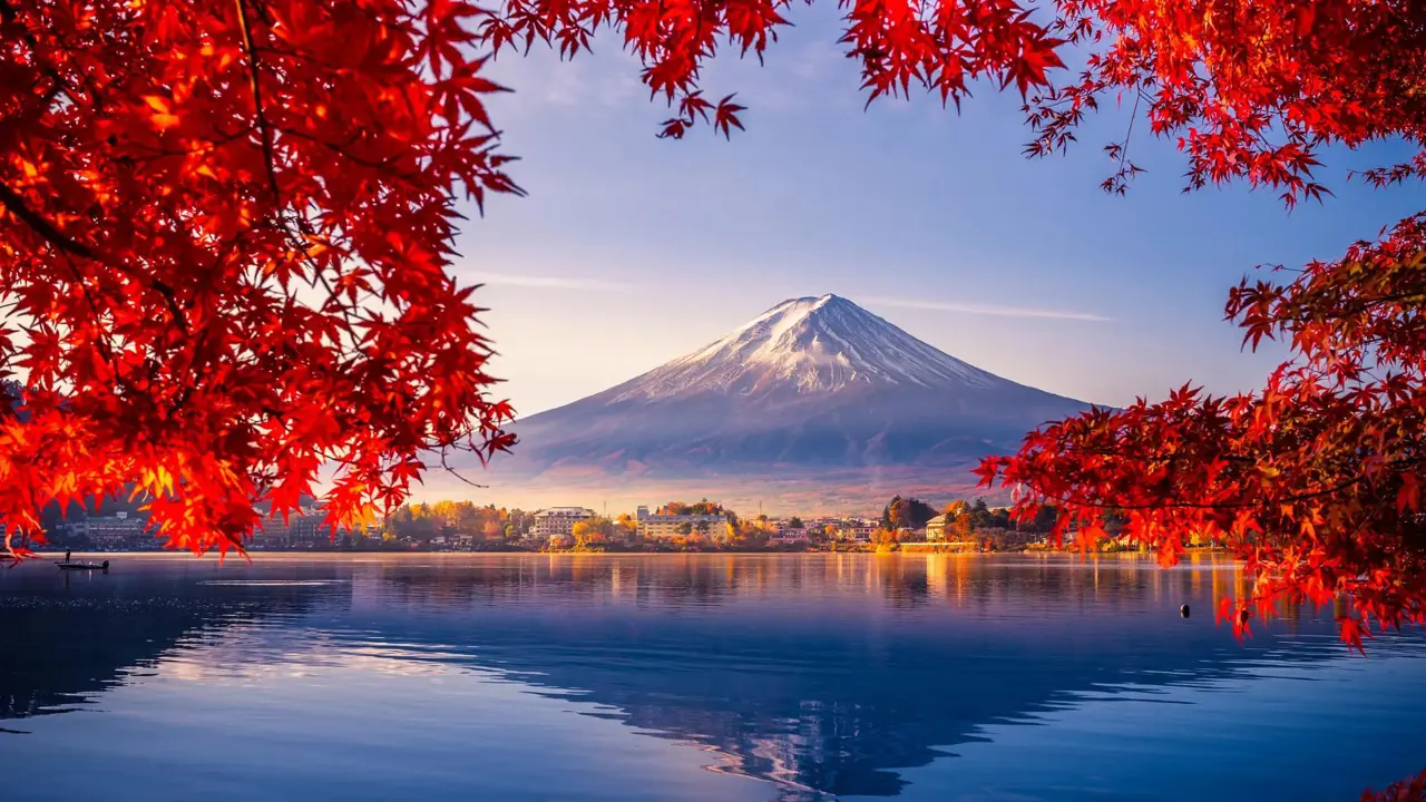 Mount Fuji reflected in Lake Kawaguchi, framed by vivid red autumn leaves, with a sunlit lakeside town between the water and the mountain